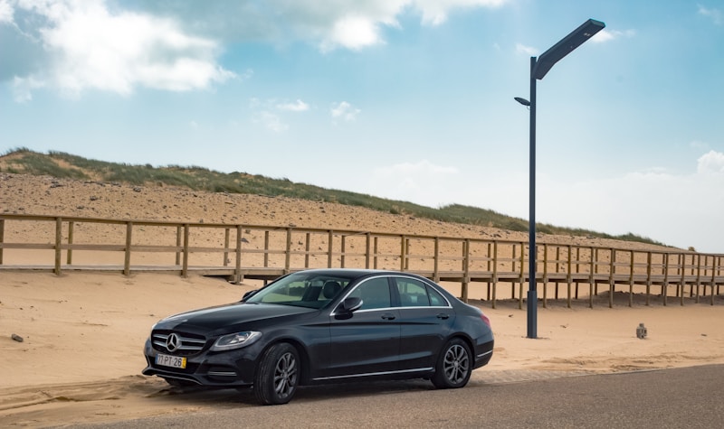 A black Mercedes-Benz sedan is parked on a sandy area near a beach. Behind the car, a wooden boardwalk runs parallel to a sandy hill covered with grass. A tall, modern streetlight stands on the right side of the image, and the sky is clear with a few clouds.