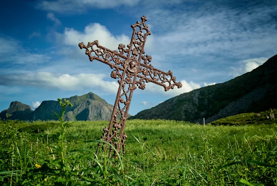 leaning brown metal cross on green grass field near mountains at daytime in Hovden Norway