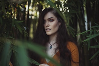 A woman with long, dark hair stands among tall green bamboo stalks, gazing into the distance. She is wearing a rust-colored top and a necklace with a dark pendant. Soft, natural light filters through the surrounding leaves, creating a serene and mysterious atmosphere.