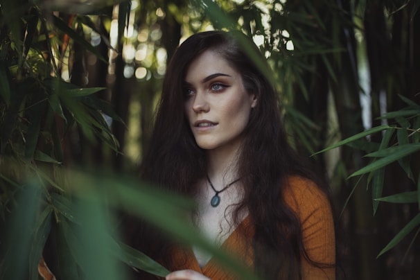 A woman with long, dark hair stands among tall green bamboo stalks, gazing into the distance. She is wearing a rust-colored top and a necklace with a dark pendant. Soft, natural light filters through the surrounding leaves, creating a serene and mysterious atmosphere.