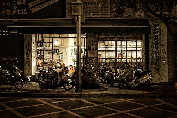 A dimly lit street scene in front of a shop with a large window showcasing various items inside. Outside, numerous scooters and motorcycles are parked on the sidewalk. The shop is illuminated, creating a contrast with the darker surroundings, and the overall setting feels quiet and still.