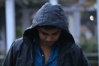 A person wearing a dark hooded jacket looks downward, creating a contemplative or introspective mood. The background is blurry, emphasizing the subject while providing context with soft colors.