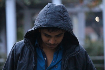 A person wearing a dark hooded jacket looks downward, creating a contemplative or introspective mood. The background is blurry, emphasizing the subject while providing context with soft colors.