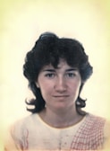 A woman is shown in a portrait, facing the camera directly with a neutral expression. Her hair is dark and curly, framing her face. She wears a light-colored top with a red patterned design on the shoulder. The background is plain and softly colored, adding focus to the subject.