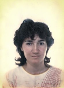 A woman is shown in a portrait, facing the camera directly with a neutral expression. Her hair is dark and curly, framing her face. She wears a light-colored top with a red patterned design on the shoulder. The background is plain and softly colored, adding focus to the subject.