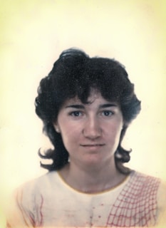 A woman is shown in a portrait, facing the camera directly with a neutral expression. Her hair is dark and curly, framing her face. She wears a light-colored top with a red patterned design on the shoulder. The background is plain and softly colored, adding focus to the subject.
