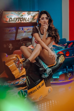 A person is sitting on an arcade motorcycle game with vibrant neon lighting. The individual wears glasses, a casual T-shirt, shorts, and boots, exuding a relaxed yet trendy vibe. The arcade setting has colorful and playful elements, enhancing the lively atmosphere.