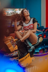 A woman wearing glasses and a shirt with the word 'SQUAD' sits on a motorcycle arcade game. She is playfully flipping her hair while seated on the yellow and black motorcycle. The background includes colorful arcade machines, and there is a relaxed and playful atmosphere.