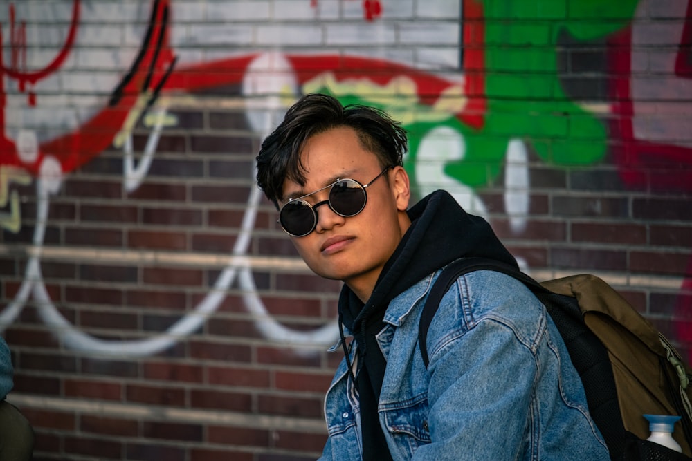 man in black sunglasses and blue denim jacket standing in front of wall