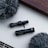 Two black miniature microphones are placed on a white surface, surrounded by two fluffy grey windshields. The background features a brown wooden edge, giving a neutral and minimalistic aesthetic.