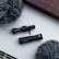 Two black miniature microphones are placed on a white surface, surrounded by two fluffy grey windshields. The background features a brown wooden edge, giving a neutral and minimalistic aesthetic.
