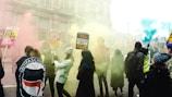 A group of people are gathered for a protest or demonstration in an urban setting, with colorful smoke around them. They hold various signs that include messages advocating for refugee rights and against racism and xenophobia. The attendees are dressed in casual clothing, and there is a mix of individuals, including some wearing hats and jackets.