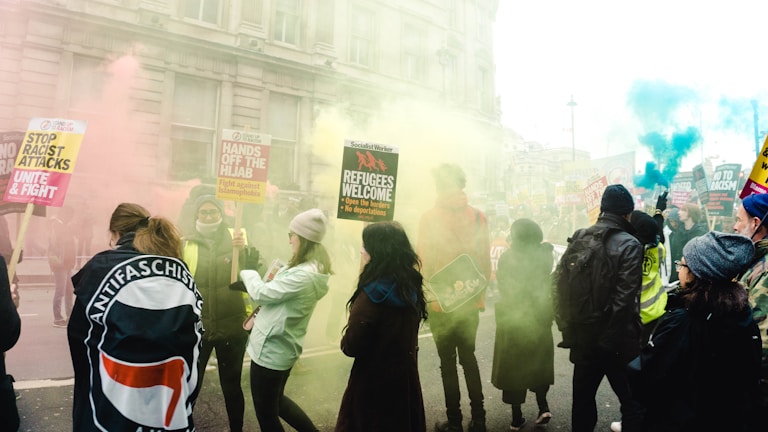 A group of people are gathered for a protest or demonstration in an urban setting, with colorful smoke around them. They hold various signs that include messages advocating for refugee rights and against racism and xenophobia. The attendees are dressed in casual clothing, and there is a mix of individuals, including some wearing hats and jackets.