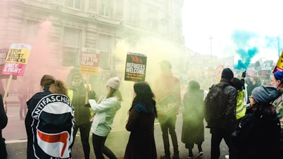A group of people are gathered for a protest or demonstration in an urban setting, with colorful smoke around them. They hold various signs that include messages advocating for refugee rights and against racism and xenophobia. The attendees are dressed in casual clothing, and there is a mix of individuals, including some wearing hats and jackets.