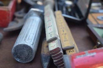 A close-up view of various hand tools and measuring devices, including a folded ruler with visible numerical markings and a large metallic tool with a cylindrical grip. The tools are arranged casually on a table, suggesting a workshop or garage setting.
