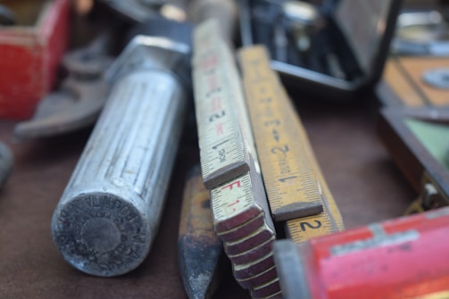 A close-up view of various hand tools and measuring devices, including a folded ruler with visible numerical markings and a large metallic tool with a cylindrical grip. The tools are arranged casually on a table, suggesting a workshop or garage setting.
