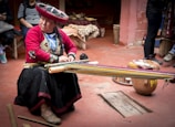 A person wearing traditional Andean clothing is seated on the floor, working on a loom. The colorful attire includes a large embroidered hat and detailed fabrics. The setting appears to be a rustic room with clay floors and wooden benches, suggesting a craft area or workshop.