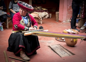 A person wearing traditional Andean clothing is seated on the floor, working on a loom. The colorful attire includes a large embroidered hat and detailed fabrics. The setting appears to be a rustic room with clay floors and wooden benches, suggesting a craft area or workshop.