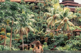 A person is relaxing in an infinity pool facing a verdant tropical landscape. The scene is dominated by lush greenery, including palm trees and dense foliage, with a traditional-style building in the background.