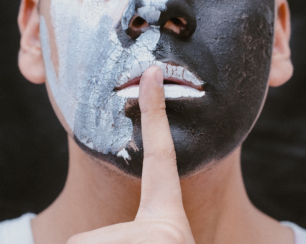 person with black and white face paint with finger on mouth doing the quiet gesture