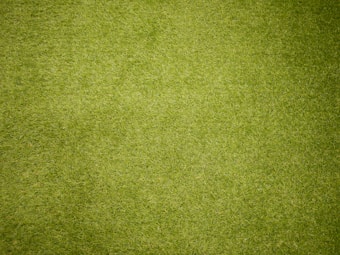 A close-up view of a green grassy surface, densely covered with neatly trimmed blades of grass. The texture is uniform, indicating well-maintained lawn or artificial turf.