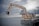 A large industrial excavator is positioned on a floating platform on a body of water. The excavator has a long articulated arm with a claw-like bucket, and a cabin for the operator. The platform appears to be a barge, indicating use for dredging or construction in aquatic environments. The sky is overcast, casting a muted light over the scene.