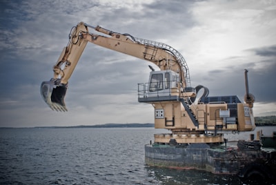 A large industrial excavator is positioned on a floating platform on a body of water. The excavator has a long articulated arm with a claw-like bucket, and a cabin for the operator. The platform appears to be a barge, indicating use for dredging or construction in aquatic environments. The sky is overcast, casting a muted light over the scene.