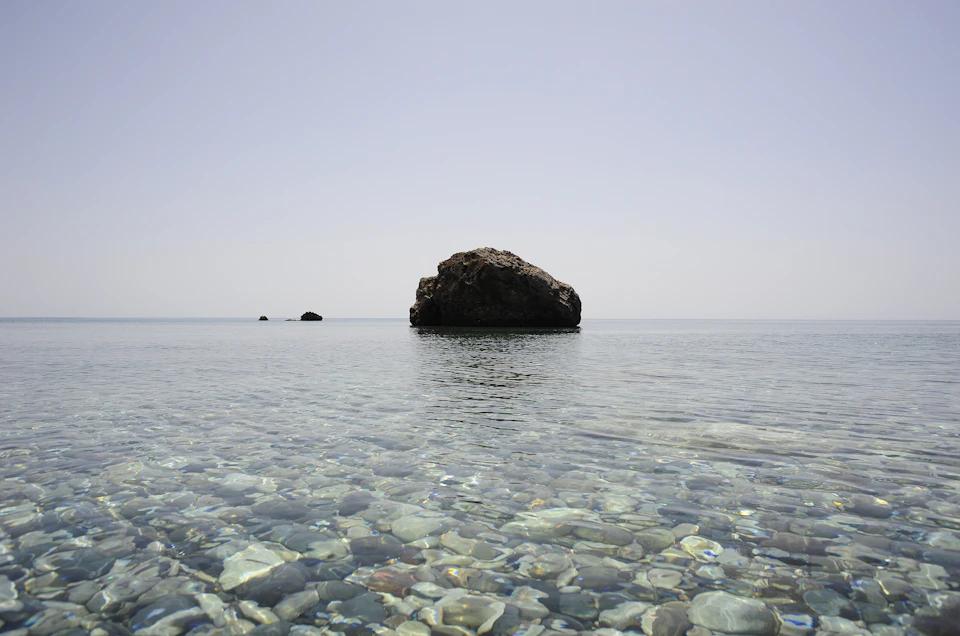 The image depicts a serene scene at sea, with two large rocks situated on the water's surface. The rock on the left is slightly larger than the one on the right, creating an interesting visual balance within the frame. Both rocks are surrounded by clear blue water that reflects the light from above, adding depth and dimension to the image. In the background, a few small islands can be seen, providing a sense of scale and context for this picturesque scene.