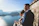 A couple, dressed in formal attire, stands by a stone wall overlooking a large, serene lake with mountainous scenery in the background. The man is wearing a suit, and the woman, possibly in a bridal dress, holds a bouquet of flowers. The view includes a small island with a building in the middle of the lake, with clear blue skies adding to the tranquil ambiance.