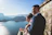 A couple, dressed in formal attire, stands by a stone wall overlooking a large, serene lake with mountainous scenery in the background. The man is wearing a suit, and the woman, possibly in a bridal dress, holds a bouquet of flowers. The view includes a small island with a building in the middle of the lake, with clear blue skies adding to the tranquil ambiance.