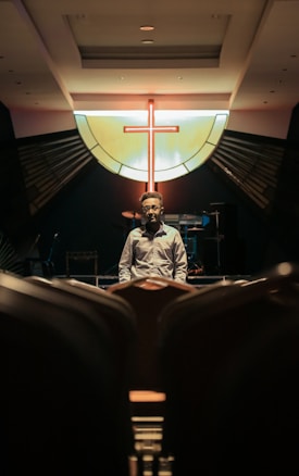 A person stands in an empty auditorium facing the camera, with a prominent backlit cross symbol on a stained glass window in the background. The lighting casts dramatic shadows, emphasizing a somber and contemplative atmosphere.
