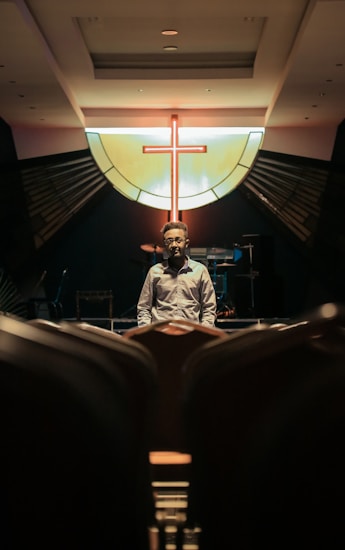 A person stands in an empty auditorium facing the camera, with a prominent backlit cross symbol on a stained glass window in the background. The lighting casts dramatic shadows, emphasizing a somber and contemplative atmosphere.