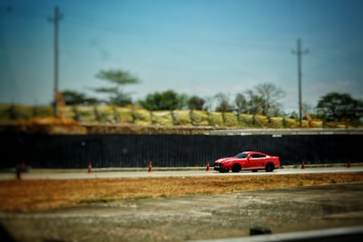 A bright red sports car is driving on a road lined with orange traffic cones. The background features a fenced area with some greenery and industrial elements. The sky is clear with a few power poles visible.