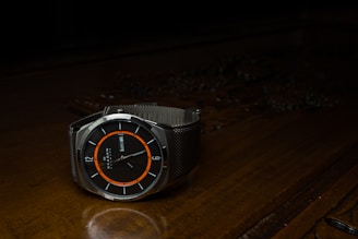 A wristwatch with a black watch face, accented by an orange ring and white markings, is resting on a dark wooden surface. The watch has a sleek, modern design with a metallic mesh band. The day and date are visibly displayed on the dial.