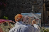 A person wearing an orange cap and white shirt is painting an urban scene on a canvas set on an easel in an outdoor setting. The painting depicts houses with red roofs, and the scene suggests a rustic or historical area. A red car is partially visible in the background, parked near a stone wall.