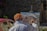 A person wearing an orange cap and white shirt is painting an urban scene on a canvas set on an easel in an outdoor setting. The painting depicts houses with red roofs, and the scene suggests a rustic or historical area. A red car is partially visible in the background, parked near a stone wall.