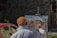 A person wearing an orange cap and white shirt is painting an urban scene on a canvas set on an easel in an outdoor setting. The painting depicts houses with red roofs, and the scene suggests a rustic or historical area. A red car is partially visible in the background, parked near a stone wall.