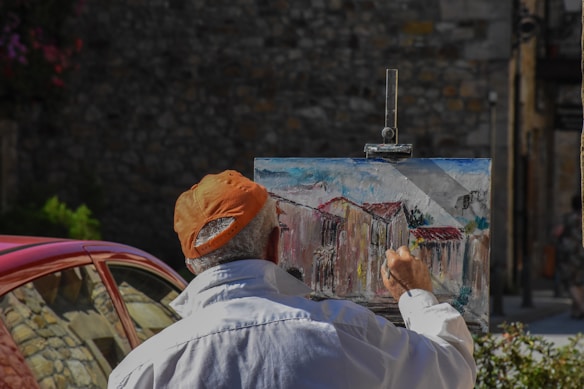 A person wearing an orange cap and white shirt is painting an urban scene on a canvas set on an easel in an outdoor setting. The painting depicts houses with red roofs, and the scene suggests a rustic or historical area. A red car is partially visible in the background, parked near a stone wall.