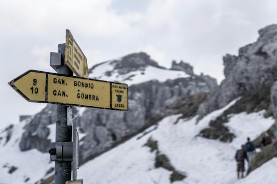 A yellow directional signpost stands in the foreground with snow-covered mountains and rocky terrain in the background. The sign indicates paths to 'C.N. Bobbio' and 'C.N. Comera' as part of the 'CAI di Lecco' trails. Hiking figures appear in the distance, and the sky is overcast.