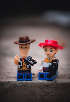 Two toy figures resembling cowboys are seated on a rough, textured surface. The figure on the left wears a brown hat and a cow-print vest, while the figure on the right has a red hat with a star. Both figures hold black objects that resemble cameras.