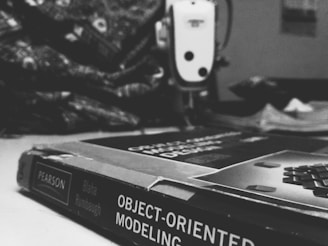 A black and white photograph of a book titled 'Object-Oriented Modeling'. The book is resting on a surface with its spine clearly showing. In the background, there is an out-of-focus sewing machine and a pile of patterned fabric. The setting seems to be a workspace, perhaps a sewing or crafting area.