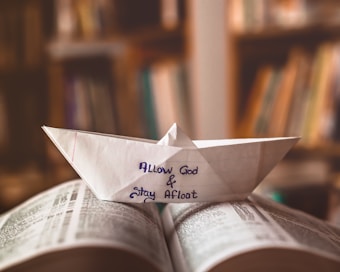 A paper boat made from a sheet of paper with the words 'Allow God & Stay Afloat' written on it is placed on top of an open book. The background is blurry and appears to show a bookshelf filled with books.