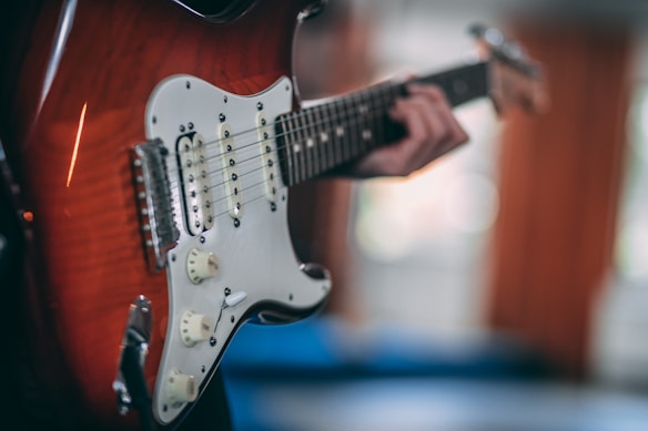 A close-up of a person playing an electric guitar, focusing on the guitar's strings and body. The guitar has a sunburst finish and a white pickguard. The background is softly blurred, giving the image a shallow depth of field.