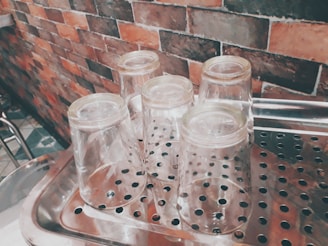Five clear, upside-down drinking glasses rest on a metal draining rack with holes. The background features a textured brick wall with shades of red, brown, and gray.