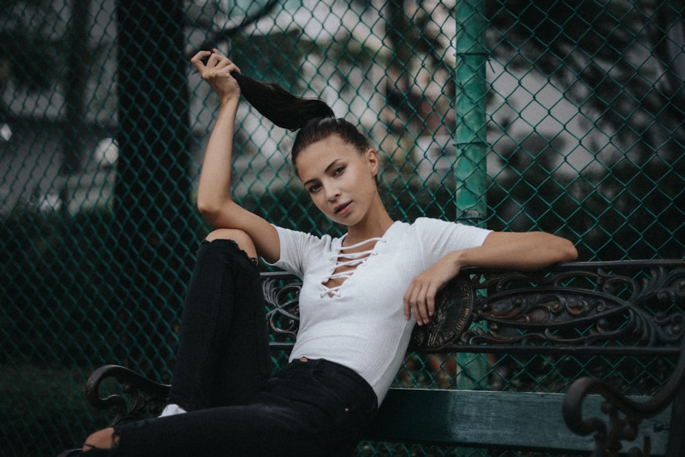 woman wearing white cap-sleeved crop top and distressed black denim jeans sitting on black metal bench beside chain-link fence during daytime