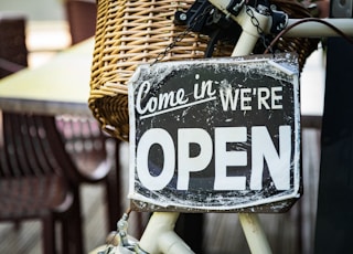 black and white come in we're open signage pinned on white bicycle with brown wicker basket at daytime