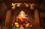 A beautifully decorated idol of Lord Ganesha is seated within an ornate altar, adorned with vibrant garlands of flowers and surrounded by intricate carvings. Soft lighting highlights the gilded details and the colorful drapery that enhances the deity's appearance.