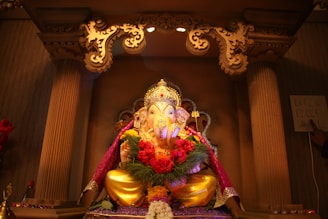 A beautifully decorated idol of Lord Ganesha is seated within an ornate altar, adorned with vibrant garlands of flowers and surrounded by intricate carvings. Soft lighting highlights the gilded details and the colorful drapery that enhances the deity's appearance.