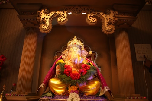 A beautifully decorated idol of Lord Ganesha is seated within an ornate altar, adorned with vibrant garlands of flowers and surrounded by intricate carvings. Soft lighting highlights the gilded details and the colorful drapery that enhances the deity's appearance.