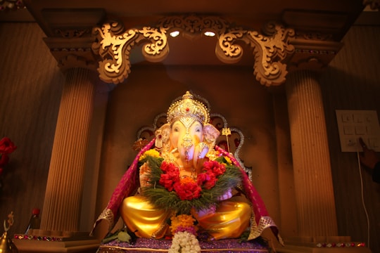 A beautifully decorated idol of Lord Ganesha is seated within an ornate altar, adorned with vibrant garlands of flowers and surrounded by intricate carvings. Soft lighting highlights the gilded details and the colorful drapery that enhances the deity's appearance.